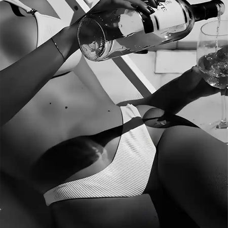 Zwart-wit beeld van een persoon die op een zonnebank ligt, een bikini draagt en een drankje uit een fles in een glas giet. De scène suggereert ontspanning en vrije tijd. Het gezicht van de persoon is niet zichtbaar.