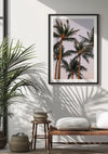 Een ingelijst schilderij Hoge Palmbomen van CollageDepot hangt aan een witte muur. Daaronder staat een geweven bank met een kussen en twee manden erop of ernaast. Natuurlijk licht filtert door een raam en werpt schaduwen van een potplant en het bankje op de muur en vloer, waardoor een serene wanddecoratie ontstaat.,Zwart