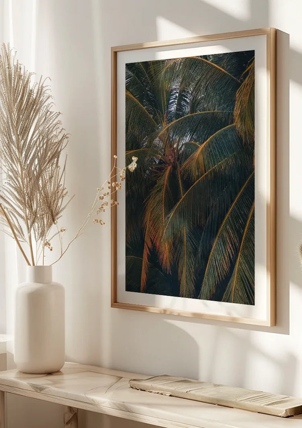 Aan een witte muur hangt een ingelijste foto van palmbladeren, die dienst doet als elegant CollageDepot Ingezoomd Op De Palmboom Schilderij. Daaronder rust een witte keramische vaas met daarin gedroogd gras en een kleine tak op een lichtgekleurde houten plank. Zonlicht verlicht het tafereel en werpt zachte schaduwen.,Lichtbruin