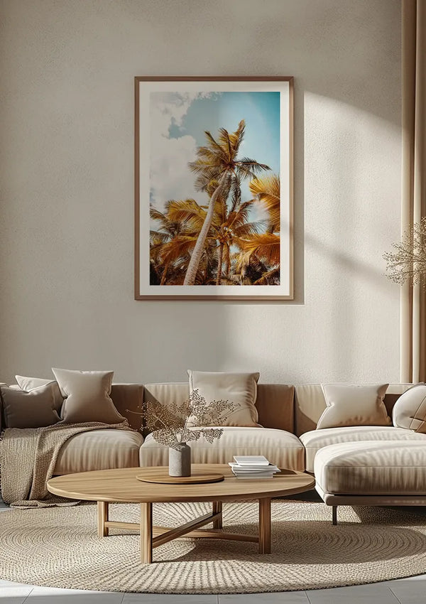Een minimalistische woonkamer heeft een grote ingelijste foto van palmbomen aan de muur, met de elegante wanddecoratie "Onderaanzicht Prachtige Palmbomen Schilderij" van CollageDepot. De kamer heeft een beige kleurenschema met een neutraal getinte bank, kussens, een ronde houten salontafel en een decor van gedroogde planten. Zachte verlichting versterkt de serene sfeer.,Lichtbruin