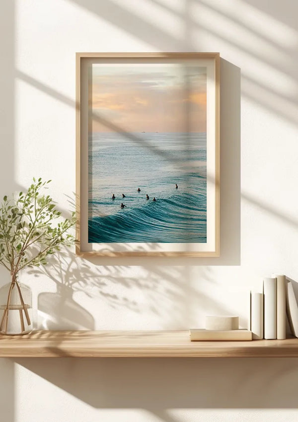 Aan een witte muur hangt een ingelijste foto van surfers in de oceaan, met behulp van een magnetisch ophangsysteem. De afbeelding legt een schilderachtig uitzicht vast met kalme golven en een zonsonderganghemel. Onder het schilderij Zwemmen In De Wilde Zee van CollageDepot staat een lichte houten plank met een plantje en een nette stapel witte boeken, perfecte wanddecoratie voor elke kamer.,Lichtbruin