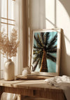 Een ingelijste Palmboom Bij Heldere Hemel Schilderij van een palmboom leunt tegen een muur op een houten tafel. Op de tafel staat ook een witte vaas met gedroogde bloemen, een kleinere witte kan en twee gestapelde boeken. Zonlicht filtert door een raam met gordijnen. Deze elegante CollageDepot wanddecoratie voegt charme toe aan de gezellige sfeer.,Lichtbruin