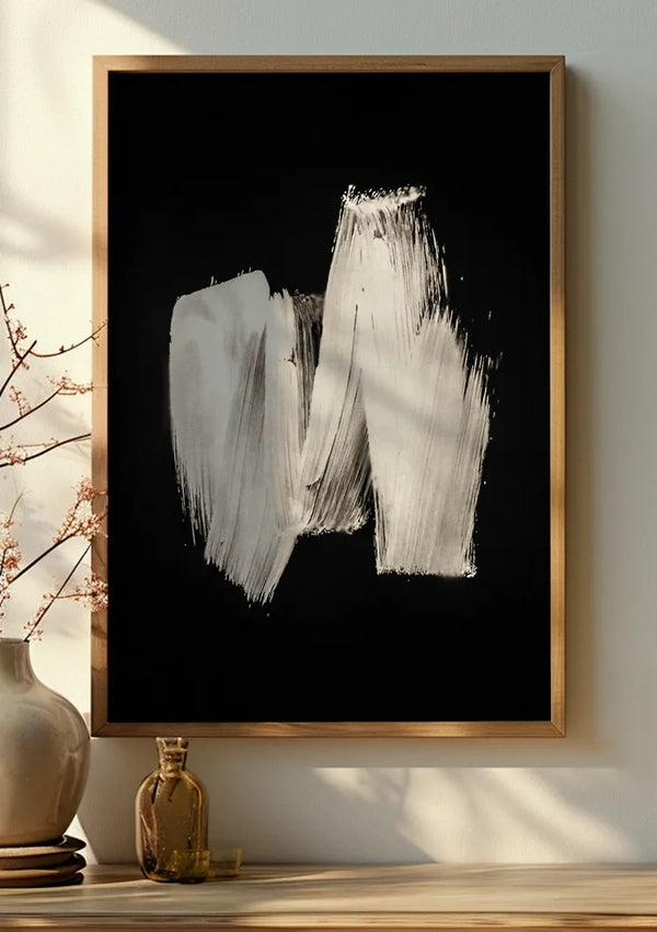 Een ingelijst Witte verfstreep schilderij van CollageDepot met brede, witte penseelstreken op een zwarte achtergrond hangt aan de muur met een magnetisch ophangsysteem. Ernaast staat een kleine vaas met lichtroze bloemen en nog een klein bruin flesje op een houten oppervlak, verlicht door zonlicht.