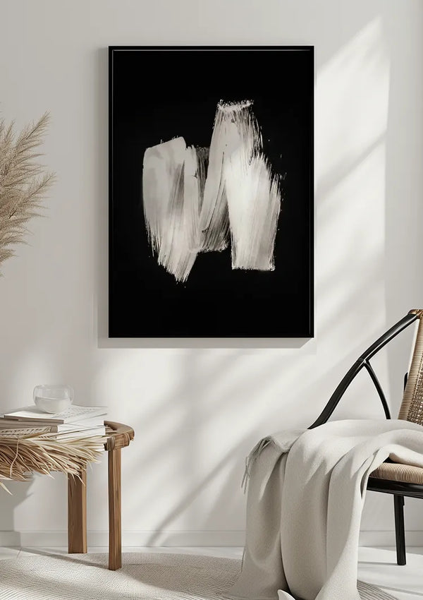 Een minimalistische kamer met een ingelijst abstract schilderij met witte penseelstreken op een zwarte achtergrond. De setting omvat een bijzettafel met boeken en een glas, een rieten stoel gedrapeerd met een witte deken en een gedroogde plant in de hoek, allemaal geaccentueerd door het ingetogen Witte verfstreep-schilderij van CollageDepot.