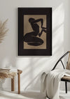 Een minimalistische kamer met een ingelijst abstract kunstwerk van een liggende figuur in de kleuren zwart en donkerbruin. De kamer is voorzien van een rieten stoel met een witte deken, een kleine houten tafel met gestapelde boeken en een glas, en pampagras. Het Abstract getekende vrouw schilderij van CollageDepot geeft een elegante toets aan de ruimte.