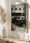 Een ingelijste spiegel of glazen paneel weerspiegelt een buitenscène met een klassieke zwarte auto geparkeerd nabij een kettingversperring. Op de achtergrond staat een wit gebouw met een bord met de tekst "Lodewijk XV Alain Ducasse." Binnen is links een vaas met gedroogde planten zichtbaar. Dit elegante Klassieke Bentley Auto Schilderij van CollageDepot voegt charme toe aan elke ruimte.,Lichtbruin