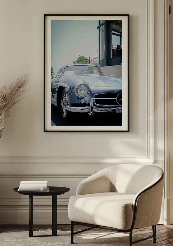 Een ingelijst CollageDepot Mercedes 300SL Schilderij hangt aan een muur boven een witte, moderne fauteuil en een klein zwart bijzettafeltje in een minimalistische woonkamer. Natuurlijk licht stroomt van rechts naar binnen en creëert zachte schaduwen op de lichtgekleurde muur en vloer, wat deze elegante wanddecoratie perfect aanvult.,Zwart