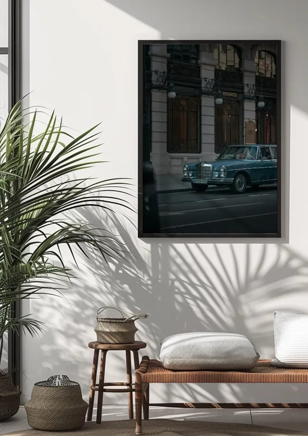 Een Blauwe Vintage Mercedes Schilderij van CollageDepot hangt aan een witte muur met behulp van een innovatief magnetisch ophangsysteem. Daaronder staat een houten bank met een kussen en een geweven mand. Links staat een hoge groene plant die een schaduw op de muur werpt: de perfecte wanddecoratie.,Zwart