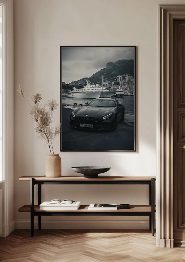 Aan een witte muur hangt een ingelijste foto van een zwarte auto die geparkeerd staat bij een jachthaven in Monaco. Hieronder staat een zwarte consoletafel met een vaas met gedroogde bloemen, een open boek en een ondiepe schaal, waardoor een minimalistische en elegante wanddecoratie ontstaat met het Mercedes AMG GTR Monaco Schilderij van CollageDepot.,Zwart