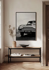 Een ingelijste zwart-witfoto van een klassieke Aston Martin uit de serie "Iconische Aston Martin Schilderij" van CollageDepot wordt aan een lichtgekleurde muur gehangen en dient als elegante wanddecoratie boven een houten consoletafel. Op de tafel staat een vaas met gedroogde planten, een open boek en een ondiepe keramische kom. De vloer is voorzien van een visgraat houtpatroon.,Zwart