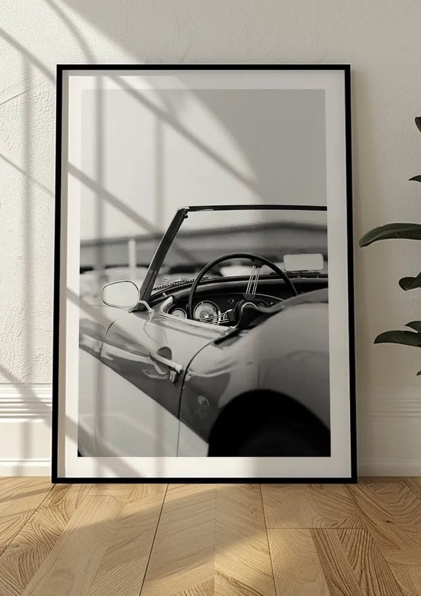 Een ingelijst zwart-wit Auto Interieur Schilderij van een vintage cabriolet van CollageDepot leunt tegen een witte muur. Het stuur en het dashboard zijn zichtbaar, met lichte schaduwen van een nabijgelegen raam die over de foto vallen. Aan de rechterkant is gedeeltelijk een potplant zichtbaar, wat charme toevoegt aan deze magnetische wanddecoratie.,Zwart