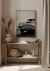 Een minimalistische kamer is voorzien van een iconisch Aston Martin Schilderij van CollageDepot, een ingelijste zwart-witfoto van een klassieke auto met een '403 GBC'-kentekenplaat, hangend boven een smalle houten consoletafel. Op de tafel staan decoratieve voorwerpen, waaronder vazen, schalen en een rieten mand, naast een beige gordijn.,Lichtbruin