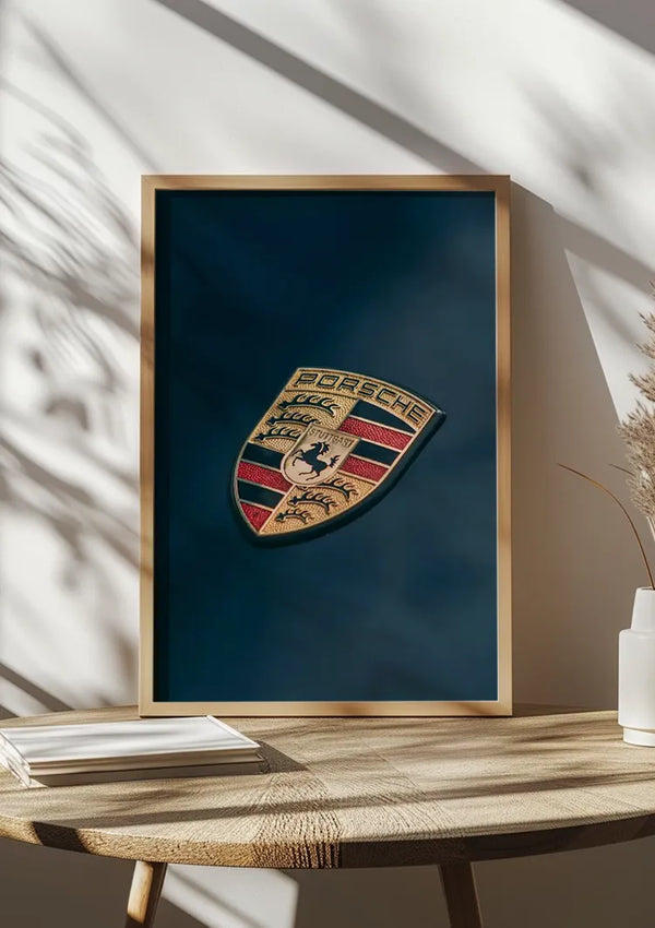 Een ingelijste CollageDepot Porsche Logo Schilderij wordt tentoongesteld op een houten tafel naast een kleine stapel gesloten boeken. De tafel, die tegen een muur staat, toont schaduwen van nabijgelegen planten die patronen op het oppervlak werpen, wat de esthetische wanddecoratie versterkt.,Lichtbruin