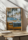 Een ingelijst CollageDepot Gele Rolls Royce Phantom Schilderij, geparkeerd voor palmbomen, tentoongesteld op een houten tafel. De scène is helder verlicht en zonlicht werpt schaduwen op de muur en de tafel. Ook liggen er wat gevouwen papieren en een sierplant op tafel.,Lichtbruin