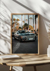 Op een houten oppervlak wordt een ingelijste foto van een Bentley-cabriolet weergegeven. De afbeelding toont de auto in een straat vol hoge palmbomen, met een helderblauwe lucht op de achtergrond. Op de tafel staat ook een kleine stapel boeken, waardoor dit elegante, prachtige Bentley-schilderij van CollageDepot perfect is voor elke ruimte.,Lichtbruin