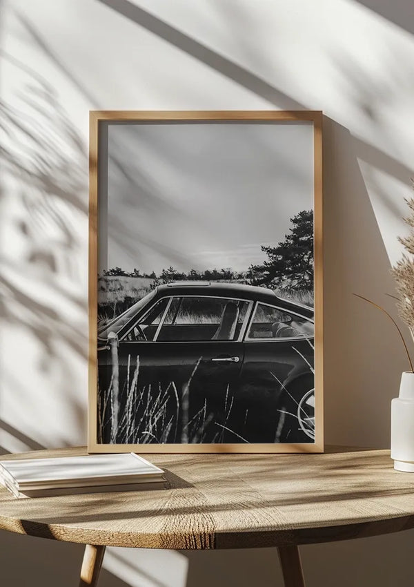 Ingelijste zwart-wit foto van een vintage auto gedeeltelijk verborgen door hoog gras, geplaatst op een houten tafel naast een witte vaas en gestapelde boeken. Zonlicht werpt schaduwen op de muur achter deze prachtige wanddecoratie, het Porsche Nature Schilderij van CollageDepot.,Lichtbruin