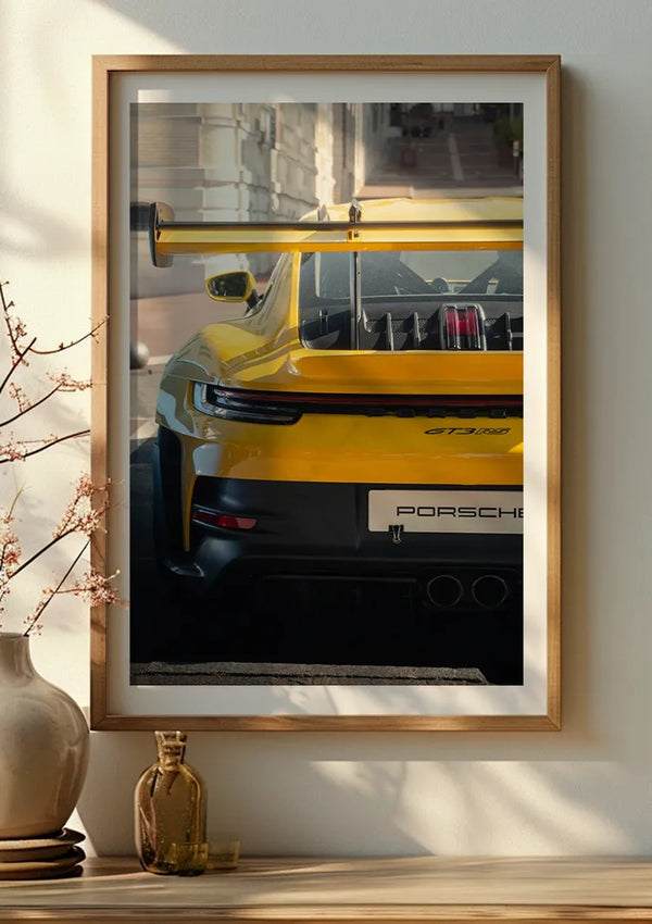 Een ingelijst geel Porsche GT3 RS-schilderij van CollageDepot wordt aan een beige muur gehangen en laat het prachtige achteraanzicht van de gele sportwagen zien. De wanddecoratie wordt aangevuld met een klein vaasje met een plant en nog een klein vaasje op een houten oppervlak onder de foto, alles beveiligd met een magnetisch ophangsysteem.,Lichtbruin