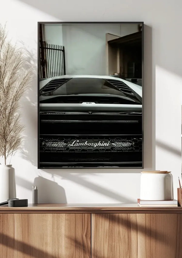 Een ingelijst Back View Lamborghini Huracan Schilderij van CollageDepot is op een witte muur gemonteerd met behulp van een magnetisch ophangsysteem. Onder de foto staat een houten plank met diverse decoratieve voorwerpen, waaronder een witte vaas met gedroogd pampagras, een witte keramische pot en een klein houten bakje.,Zwart