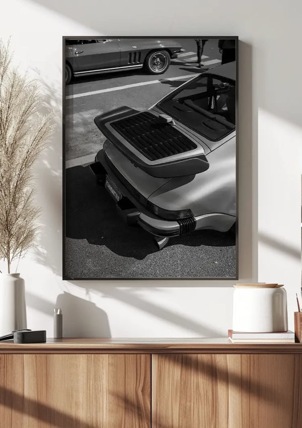 Een zwart-witte wanddecoratie van een vintage Porsche schilderij met een open motorruimte achterin is ingelijst en hangt aan een witte muur. De foto bevindt zich boven een houten kast. Op de kast staat hoog gras in een witte vaas, een kleine witte pot, een boek en wat ornamenten.,Zwart