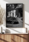 Een zwart-wit ingelijste foto van een luxe Bugatti-schilderij geparkeerd voor een groots gebouw met met klimop begroeide muren. De foto, onderdeel van een elegante wanddecoratiecollectie, hangt boven een moderne houten kast met decoratieve items, waaronder een witte vaas, een klein potje en een hoog pampagrasarrangement uit het Zwarte Bugatti Schilderij van CollageDepot.,Zwart