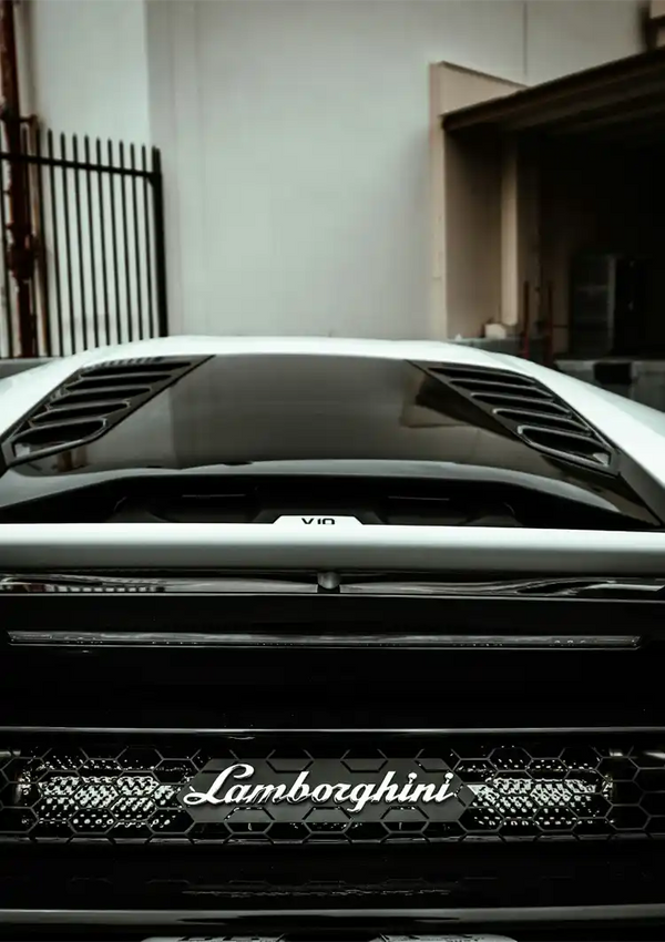 Een close-up van de achterkant van een zwarte CollageDepot aaa 013 - auto, met de nadruk op de V10-motorkap en het merklogo op de grille. De auto staat geparkeerd in een smal steegje met een hek op de achtergrond.-