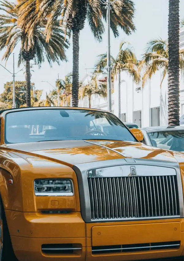 Een luxe gouden CollageDepot aaa 031 - auto's geparkeerd in een zonnige straat, omzoomd met hoge palmbomen onder een helderblauwe lucht. De kenmerkende grille en het embleem van de auto zijn duidelijk zichtbaar.-