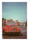Een rode vintage auto met een kentekenplaat met de tekst "SO 3 8108" staat geparkeerd op een stadsplein met geplaveide bestrating. Achter de auto bevinden zich verschillende gebouwen met klassieke architectuur en een helderblauwe lucht erboven, die doet denken aan een voortreffelijk Klassieke Rode Porsche 356 Schilderij van CollageDepot. Op de achtergrond lopen mensen.-