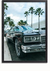 Een klassieke, glanzend zwarte Chevy Caprice met grote chromen wielen staat geparkeerd langs een straat vol palmbomen. De lucht is gedeeltelijk bewolkt. De foto lijkt ingelijst met een zwarte rand, perfect voor wanddecoratie. Deze prachtige afbeelding is verkrijgbaar als het Chevy Caprice Schilderij van CollageDepot.