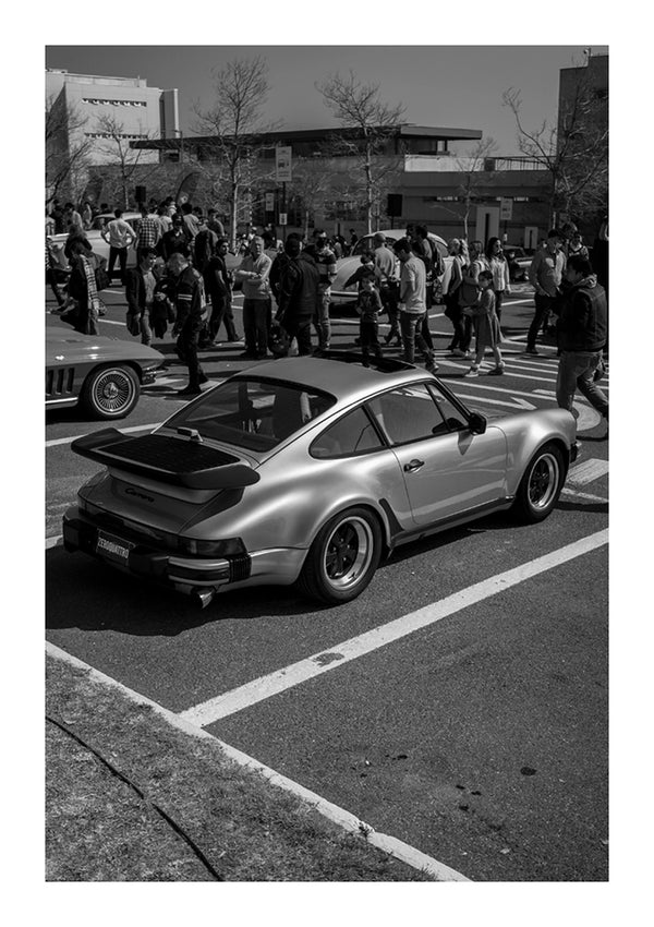 Een vintage zilveren CollageDepot Porsche aaa 075 met een grote achterspoiler geparkeerd op straat tijdens een autoshow, met groepen mensen op de achtergrond.-