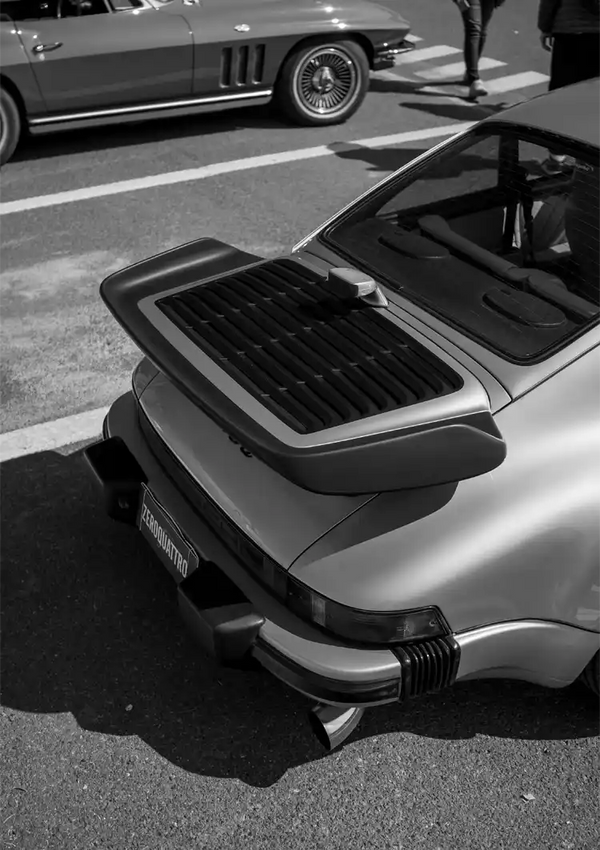 Een zwart-witfoto van het achteraanzicht van een vintage CollageDepot aaa 077 - auto's, met een verhoogde achterspoiler, geparkeerd in een zonovergoten straat naast een andere klassieke auto.-