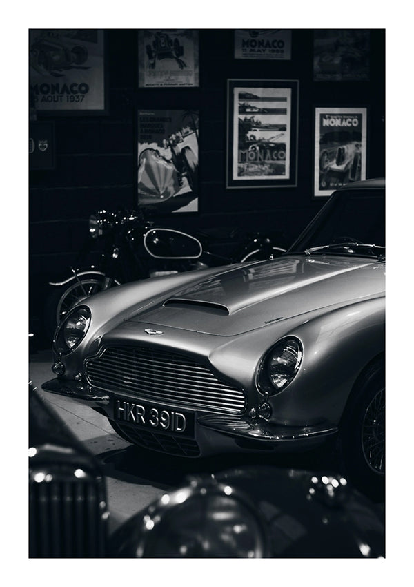 Een klassiek zilveren CollageDepot aaa 080 - auto geparkeerd in een slecht verlichte garage, met vintage Grand Prix-posters van Monaco aan de muren en een motorfiets op de achtergrond.-