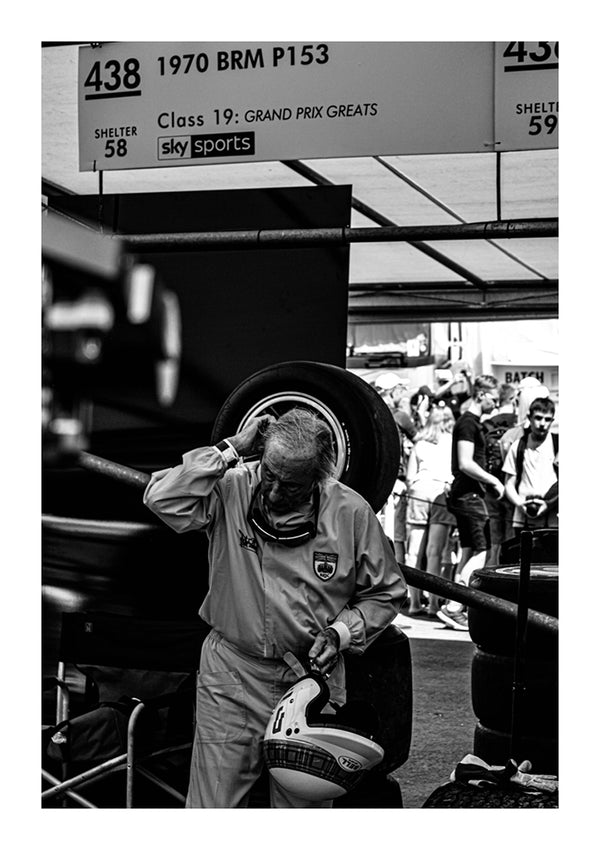 Een oudere man in een race-uniform past zijn helm aan bij een CollageDepot aaa 082 - een Formule 1-auto van een auto in een druk paddockgebied, met banden en toeschouwers zichtbaar op de achtergrond.-