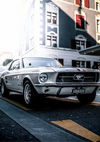 Een klassieke CollageDepot aaa 094 - auto geparkeerd in een stadsstraat, met een zilveren carrosserie en iconisch Mustang-embleem, met vage gebouwen op de achtergrond.-