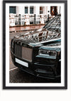 Close-up van de voorkant van een zwarte luxeauto met een prominente grille en strakke koplampen, geparkeerd op een natte stadsstraat. De auto weerspiegelt de omliggende gebouwen en het trottoir en lijkt op een verfijnde Glanzende Zwarte Rolls Royce Phantom Schilderij. De afbeelding is omlijst met een eenvoudige zwarte rand van CollageDepot.,Zwart-Met,Lichtbruin-Met,showOne,Met