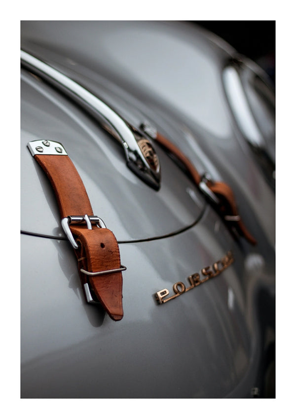 Close-up van een vintage CollageDepot aaa 114 - de grijze carrosserie van een auto met een prominent 'PORSCHE'-embleem. Kenmerken zijn onder meer bruine leren riemen met metalen gespen die de capuchon vastzetten.-