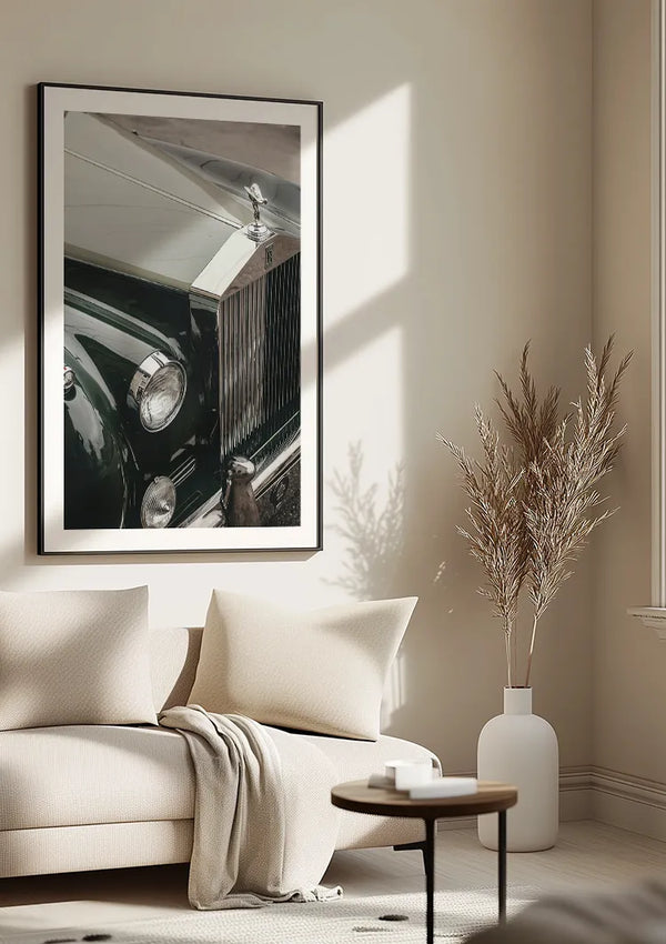Een ingelijst CollageDepot Rolls-Royce Motorkap Schilderij hangt aan een beige muur boven een lichtgekleurde bank, op zijn plaats gehouden door een magnetisch ophangsysteem. Naast de bank staat een kleine ronde tafel met een vaas met gedroogd gras, badend in natuurlijk zonlicht.,Zwart