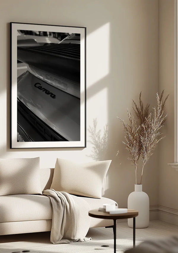 Een minimalistische woonkamer is voorzien van een beige bank met een lichte deken, een kleine ronde salontafel en een decoratieve vaas met gedroogde takken. Boven de bank hangt een groot Zwart-Wit Porsche Carrera Schilderij van CollageDepot, dat dient als opvallende wanddecoratie.,Zwart
