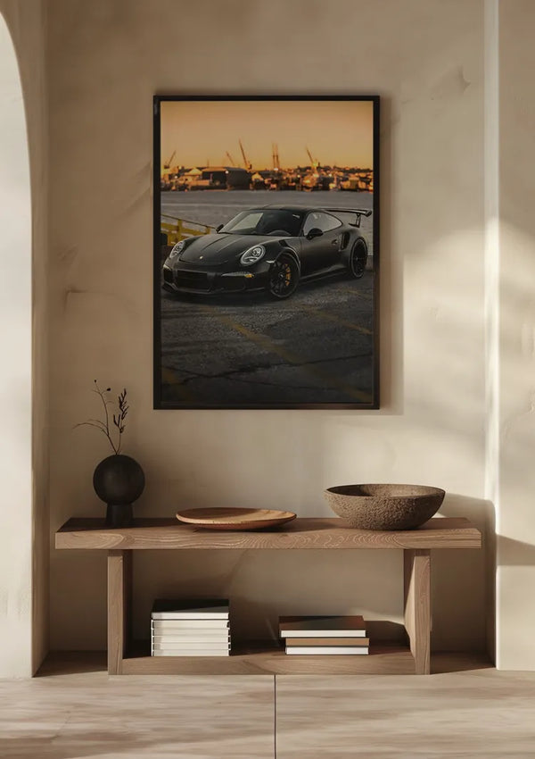 Aan een beige muur boven een houten consoletafel hangt een CollageDepot Mat Zwarte Porsche GT3 RS Schilderij met een zwarte sportwagen. Op de tafel staan twee boeken, een ronde vaas met een plant, een sierbord en een kom. Zonlicht werpt schaduwen op de muur en tafel, waardoor een elegante wanddecoratie ontstaat.,Zwart