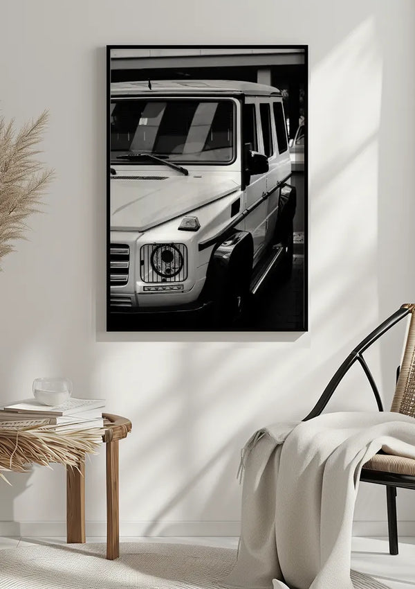 Een minimalistische kamer met een zwart-wit foto van een witte Mercedes G wagon Schilderij van CollageDepot als wanddecoratie aan de muur. Onder het schilderij bevindt zich een rieten bijzettafel met een boek en decorartikelen. Naast de tafel staat een rieten stoel met een witte deken eroverheen gedrapeerd.,Zwart
