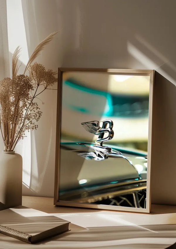 Een Prachtig Bentley Embleem Schilderij van CollageDepot staat op een houten oppervlak. De foto toont een close-up van een metalen voorwerp met een Bentley-embleem. Naast de ingelijste foto staat een vaas met daarin droogbloemen. Zacht zonlicht stroomt door een raam en verlicht het wanddecoratietafereel.,Lichtbruin