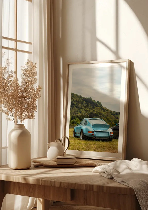 Een ingelijst Babyblauw Porsche 911 Schilderij van CollageDepot staat op een houten tafel bij een zonovergoten raam. Op de tafel staat ook een keramieken vaas met gedroogde bloemen, een kleinere vaas, een gesloten boek en een lichtgekleurd kleed. Het tafereel straalt een zachte, warme sfeer uit, perfect voor stijlvolle wanddecoratie.,Lichtbruin
