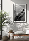 Aan een witte muur hangt een ingelijst Relaxed baden schilderij van CollageDepot van een zwemmende persoon. Onder het frame staat een houten bankje met een kussen en twee geweven manden. Links staat een potplant die schaduwen op de muur en de vloer werpt, wat de serene wanddecoratie versterkt.
