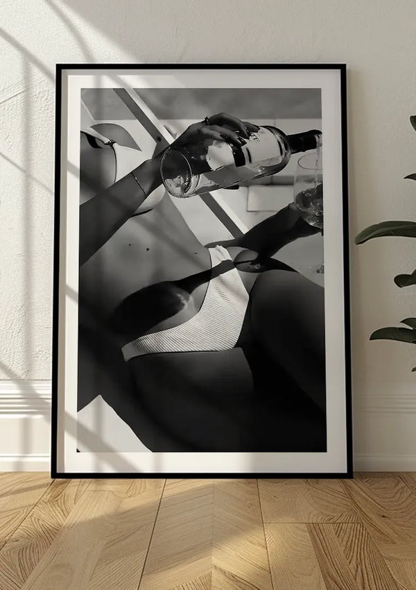 Een luxe wanddecoratie met een zwart-wit schilderij van een persoon in badkleding die een drankje uit een fles in een glas schenkt. De ingelijste foto, Summer Tanning Schilderij van CollageDepot, rust tegen een muur op een houten vloer, met schaduwen van het raam eroverheen. Rechts is gedeeltelijk een plant zichtbaar.,Zwart