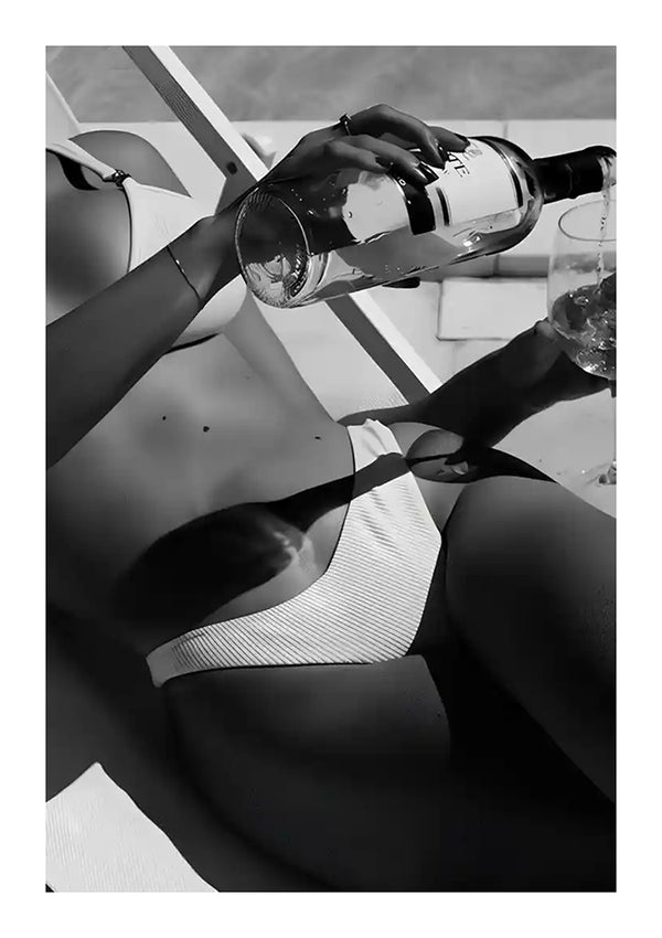 Een persoon in bikini ligt op een luie stoel en giet een drankje uit een fles in een glas. De afbeelding, die doet denken aan luxe wanddecoratie, is in zwart-wit. De setting lijkt buiten te zijn, mogelijk bij een zwembad of op het strand, waardoor de perfecte scène ontstaat voor een elegant CollageDepot Summer Tanning Schilderij.-