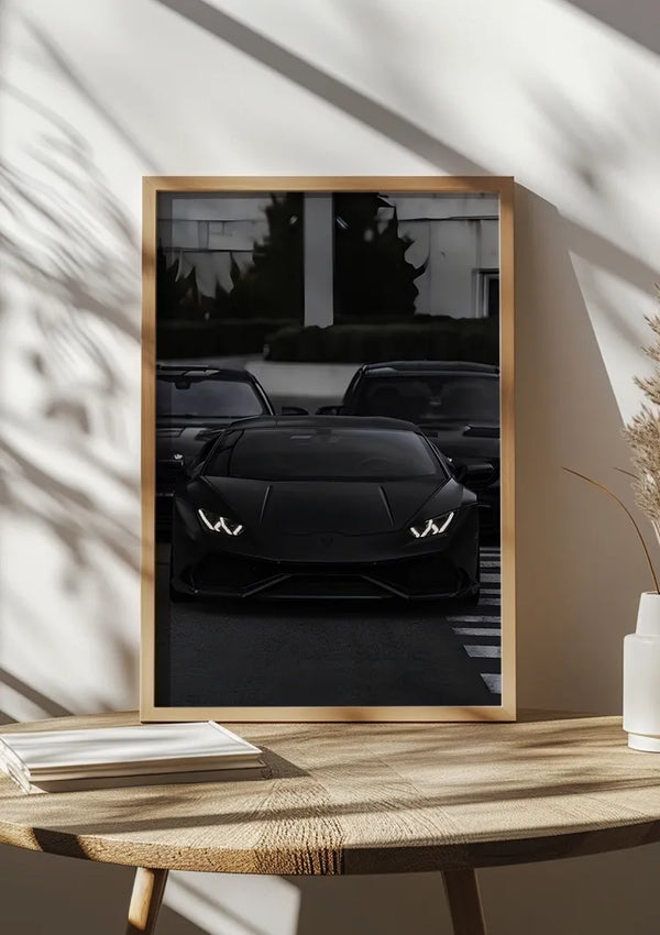 Een ingelijst zwart op zwart Lamborghini-schilderij van CollageDepot wordt weergegeven op een houten oppervlak. De koplampen van de auto branden en de auto staat geparkeerd tussen twee andere voertuigen. De foto wordt verlicht door natuurlijk daglicht dat naar binnen stroomt en schaduwen op het oppervlak werpt. Decoratieve objecten zijn dichtbij en versterken deze unieke wanddecoratie.,Lichtbruin