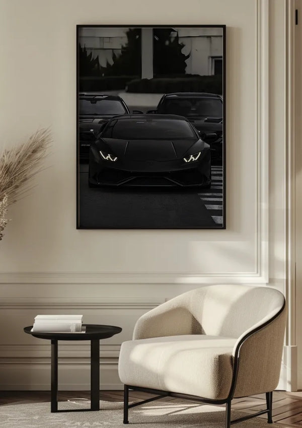 Een Zwart op Zwart Lamborghini Schilderij van CollageDepot hangt aan een beige muur boven een moderne, crèmekleurige fauteuil en een kleine, ronde zwarte tafel. De scène speelt zich af in een minimalistische woonkamer met een elegante inrichting, waaronder een vaas met gedroogd blad.,Zwart