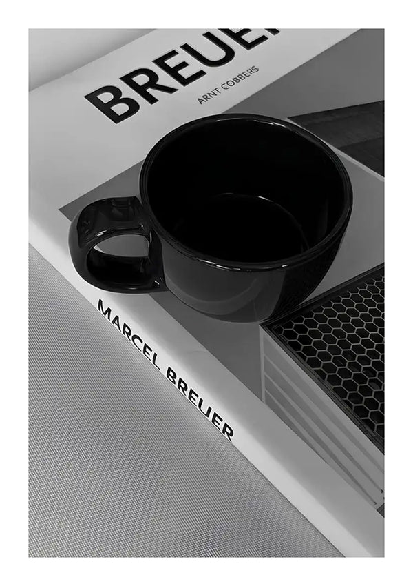 Een zwarte koffiemok van CollageDepot staat bovenop een boek met de titel "Marcel Breuer" in een monochrome omgeving, met de nadruk op minimalistische designelementen. De afbeelding is scherp, met een hoog contrast tussen de mok, de tekst en de achtergrond met patroon.-