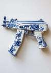 Een CollageDepot aab 332 Delfts blauw met een sierlijk blauw en wit bloemmotief op het oppervlak. Het ingewikkelde patroon lijkt op traditionele porseleinkunst en contrasteert met de vorm van het wapen. De achtergrond is effen en gebroken wit.-