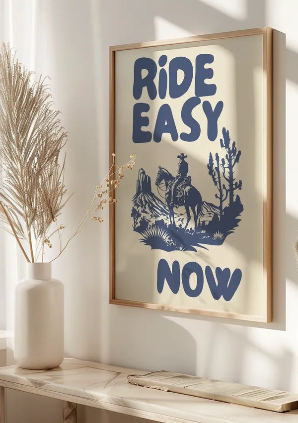 Aan de muur hangt een ingelijst "Ride Easy Now Schilderij" van CollageDepot, met de tekst "RIDE EASY NOW" in opvallende blauwe letters. Het ontwerp bevat een illustratie van een cowboy die op een paard rijdt, omringd door woestijnlandschap met cactussen. Naast het frame op een tafel staan decoratieve planten in witte vazen, wat bijdraagt aan de westers landschap wanddecoratie.,Lichtbruin