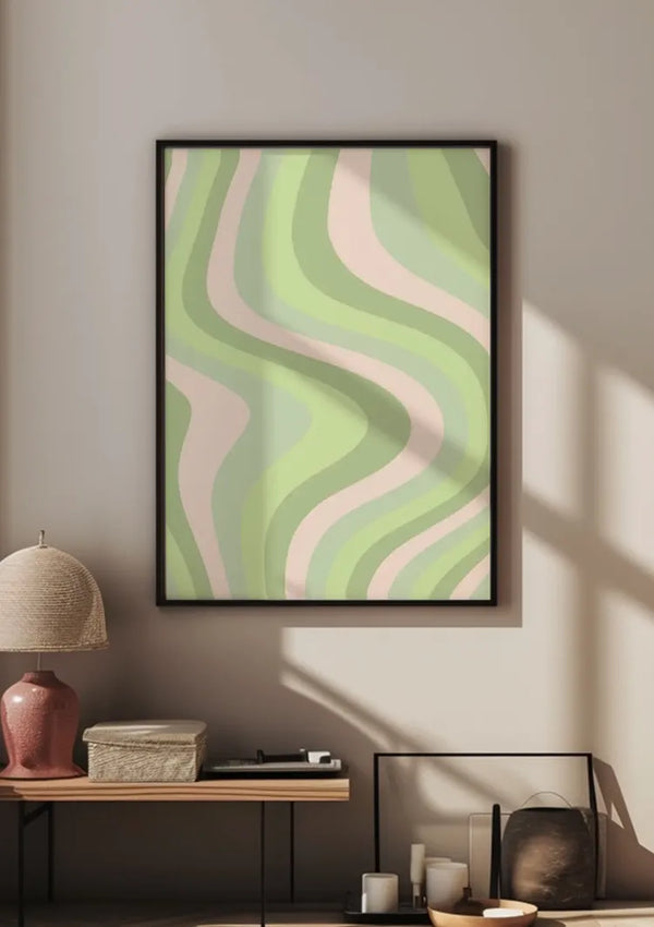 Een ingelijst goudkleurig patroon groen schilderij van CollageDepot met golvende groene en roze lijnen is gemonteerd op een beige muur. Onder de wanddecoratie staat een modern bijzettafeltje met daarin een geweven lampenkap, een stapel boeken en een glazen container. Kaarsen en decoratieve voorwerpen staan vlakbij op de vloer.